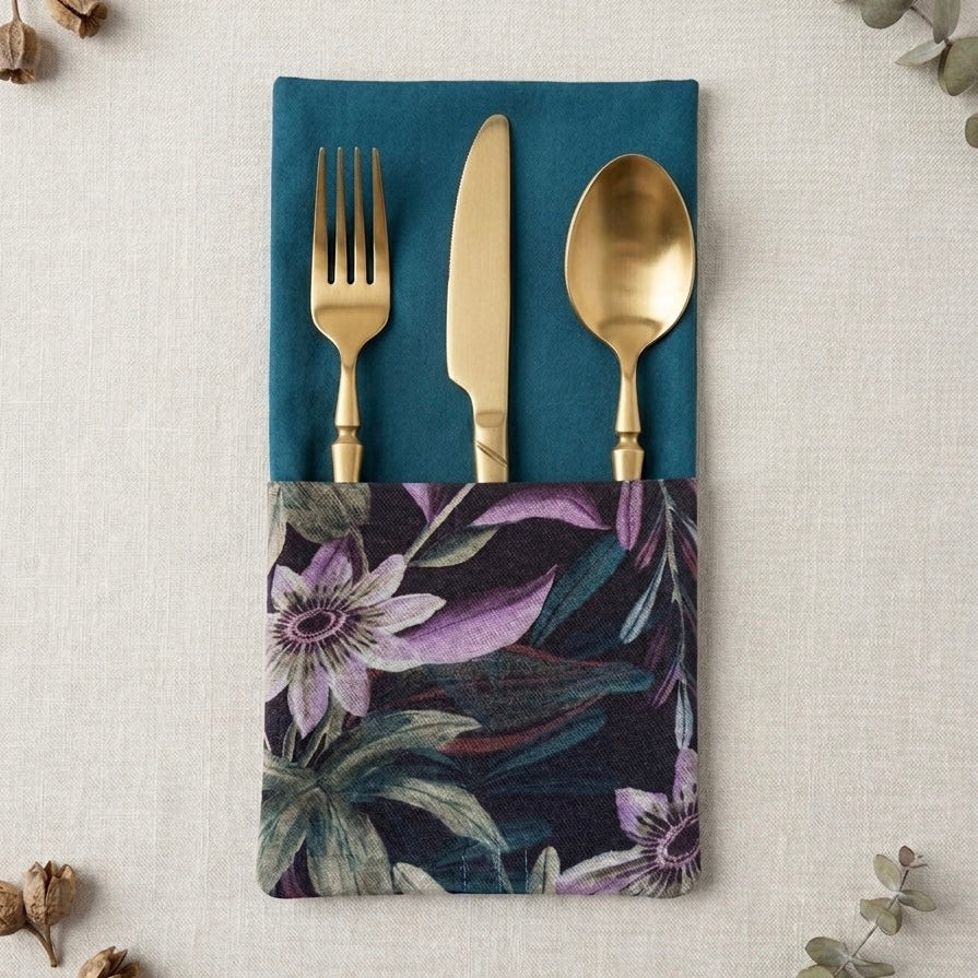servilleta de algodón en azul petroleo con bolsillo estampado de flores passifloras en tonos verdes y malvas con cubiertos dorados dentro del bolsillo